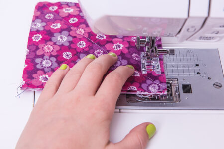 person holding a square of pink floral fabric under a sewing machine needle